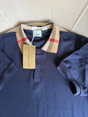 Marinblå pikétröja - Snygg marinblå pikétröja från b med klassisk beige krage i det ikoniska rutmönstret och knappar framtill. Kortärmad modell i mjukt bomullsmaterial, perfekt för en clean och stilren look. Den är i storlek M