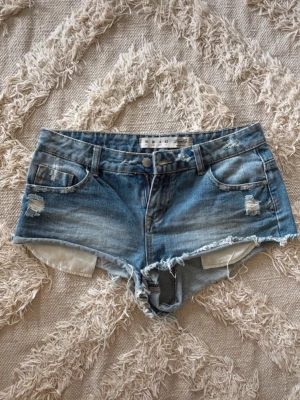 Blå lågmidjade jeansshorts med slitningar - Säljer ett par blå denimshorts med råa kanter och slitna detaljer framtill. Shortsen har  fickor, bälteshällor och knappgylf. Perfekta för en avslappnad stil och passar till det mesta. Lågmidjade storlek 36💓💓