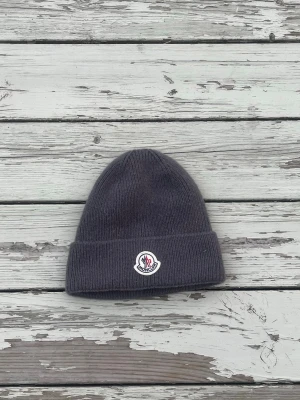Mörkgrå Moncler mössa - Säljer en stilren mörkgrå mössa från Moncler med uppvikt kant och klassisk Moncler-logga framtill. Mössan är ribbstickad och gjord i mjukt material som håller dig varm under kalla dagar. Perfekt accessoar för en trendig och enkel look.