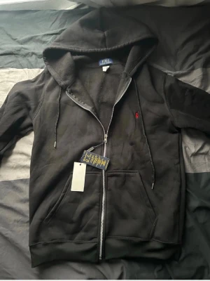 Svart hoodie från Polo Ralph Lauren - Säljer en svart hoodie med dragkedja från Polo Ralph Lauren. Klassisk design med huva, dragsnören och känguruficka framtill. Liten röd logga broderad på bröstet. Tillverkad i mjuk bomullsblandning för skön känsla.