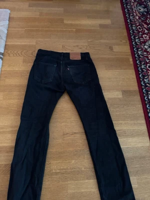 Svarta Levi's 501 jeans - Säljer ett par klassiska svarta Levi's 501 jeans med raka ben och femficksmodell. Jeansen har läderpatch bak i midjan och den ikoniska röda Levi's-taggen på bakfickan. Tillverkade i slitstark denim, perfekta för en clean och tidlös look.