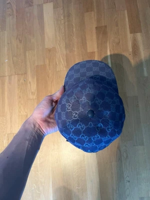 Mörkblå Gucci keps med GG-mönster - Snygg mörkblå keps från Gucci med det klassiska GG-monogrammet över hela kepsen. Justerbar baktill och insida i ljus bomull. Perfekt accessoar för dig som vill ha en exklusiv och trendig look.