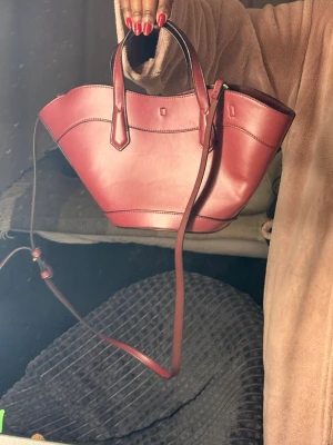Röd handväska i läder från H&M - Snygg röd handväska från H&M i slätt läder med både handtag och justerbar axelrem. Väskan har en unik form med bred öppning och fina sömdetaljer. Perfekt för dig som vill ha en trendig accessoar med plats för det viktigaste.