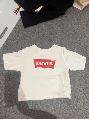 Vit Levi's t-shirt med röd logga - Säljer en klassisk vit t-shirt från Levi's med den ikoniska röda loggan tryckt på bröstet. T-shirten har korta ärmar, rund halsringning och är gjord i mjuk bomull. Perfekt för en enkel och stilren look.