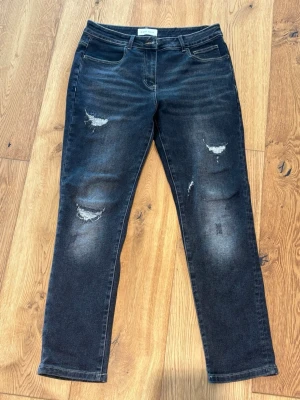 Svarta slitna jeans - Säljer ett par svarta jeans med slitningar och hål på framsidan. normal midja och raka ben. Materialet är stretchigt denim som sitter skönt och ger en avslappnad look.