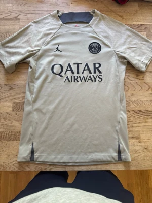 Beige PSG träningströja Jordan - Snygg beige träningströja från PSG i samarbete med Jordan. Tröjan har korta ärmar, rund hals och svart PSG-logga samt Jordan-logga på bröstet. Qatar Airways tryckt på framsidan. Tillverkad i lätt och ventilerande material, perfekt för fotbollsträning.