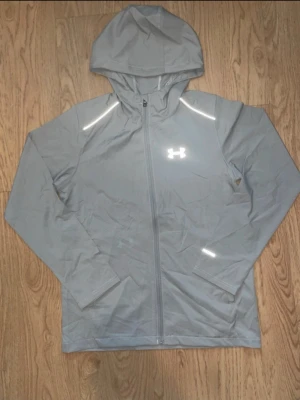 Grå träningsjacka från Under Armour - Snygg grå träningsjacka från Under Armour med hel dragkedja och huva. Jackan har reflekterande detaljer på axlar och ärm samt logga på bröstet. Tillverkad i lätt och funktionellt material som passar perfekt till löpning eller gym.