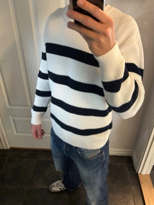 Randig tröja - Säljer en snygg stickad tröja i vitt med breda marinblå horisontella ränder. Tröjan har rund halsringning, långa ärmar och ribbade muddar. Perfekt för dig som gillar klassisk och stilren look. Materialet känns mjukt och bekvämt.