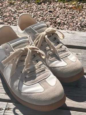 Masion margiela  - Snygga sneakers i vitt och beige med ovandel i läder och mockadetaljer. Skorna har klassisk låg profil, vita snören och en brun gummisula som ger en retrovibe. Perfekta för dig som gillar stilrena och tidlösa sneakers.