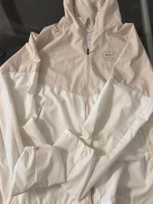 Nike Air beige och vit vindjacka - Snygg vindjacka från Nike Air i beige och vitt med dragkedja framtill och huva. Jackan har stora Nike Air-loggor både fram och bak, samt elastiska muddar vid ärmsluten. Tillverkad i lätt och vindtätt material, perfekt för blåsiga dagar.