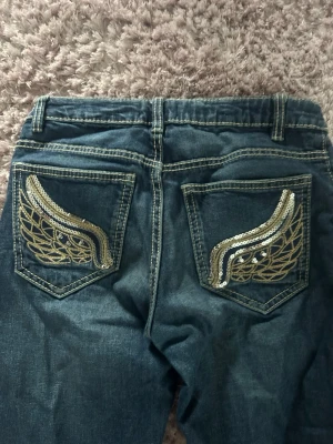 Blå bootcut jeans med broderade fickor - Säljer ett par blå bootcut jeans med snygga slitningar och kontrastsömmar. Baksidan har broderade fickor med coolt mönster i beige. Klassisk femficksmodell och låg midja. Perfekta för dig som gillar en avslappnad men trendig stil.
