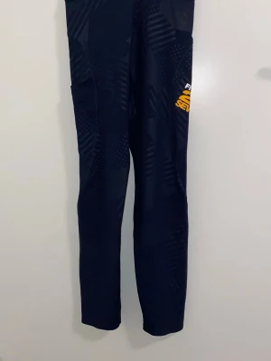 Mörkblå sportleggings  - Säljer ett par mörkblå sportleggings från Fusion med diskreta mönster och en gul logga på låret. Tight passform och stretchigt material som passar perfekt till träning eller löpning. Snygg design med detaljer som ger en sportig look.