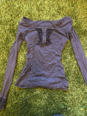 Epilouge grå henley med spetsdetaljer - Grå henley med snygga spetsdetaljer vid ärmslut och framtill. Toppen är ribbad och har u-ringning och knappar. Feminin, edgy och utstickande från mängden!
