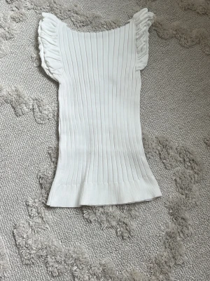 Vit ribbad topp med volangärmar - Supersöt vit ribbad topp med volangdetaljer på axlarna. Toppen är ärmlös och har en tight passform som framhäver figuren. Perfekt att styla med jeans eller kjol för en fräsch look.