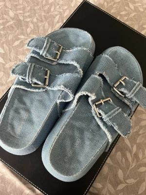 Blå denim sandaler med dubbla spännen - Trendiga sandaler i ljusblå denim med två justerbara remmar och silvriga metallspännen. Råa kanter ger en avslappnad och cool look. Perfekta för dig som gillar streetstyle och vill sticka ut med ett unikt materialval på fötterna.