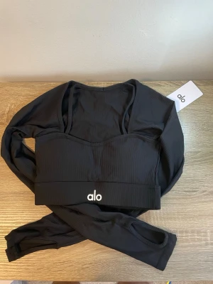 Svart långärmad topp från Alo - Snygg svart långärmad topp från Alo med diskret ribbad struktur och logga framtill. Tillverkad i stretchigt syntetmaterial som passar perfekt för träning. Tight passform och tumhål i ärmsluten för extra komfort.