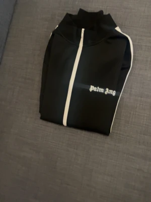 Svart zip-tröja från Palm Angels - Cool svart zip-tröja från Palm Angels med vit dragkedja och vita detaljer längs ärmarna. Tröjan har hög krage, två fickor med dragkedja och logotyp på bröstet. Perfekt för en avslappnad streetwear-look.