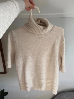 Beige stickad polotröja kort ärm - Mysig beige stickad polotröja med korta ärmar och bred ribbad kant vid hals, ärmslut och nederkant. Perfekt för lager på lager och har en avslappnad, trendig look. Fint skick