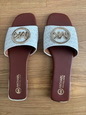 Michael Kors sandaler med logga - Snygga sandaler från Michael Kors med bred rem i ljusgrått mönster och stor guldfärgad MK-logga framtill. Innersulan är mörkbrun och har gulddetaljer. Sandalerna har fyrkantig tå och låg klack, perfekta för en trendig look.