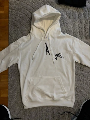 Vit hoodie - Snygg vit hoodie med svart grafiskt tryck på bröstet. Klassisk känguruficka framtill och justerbar huva med svarta snören. Perfekt för en chill och stilren look. Mjuk bomullskänsla och relaxed fit.