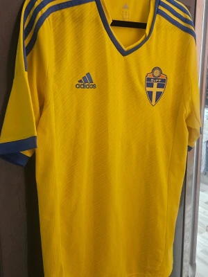 Svensk landslagströja Adidas gul - Gul svensk landslagströja från Adidas med blå detaljer och SvFF-emblem på bröstet. Tröjan har korta ärmar, blå ränder på axlarna och V-ringad krage. Perfekt för fotbollsfans som vill stötta Sverige på matchdagar.