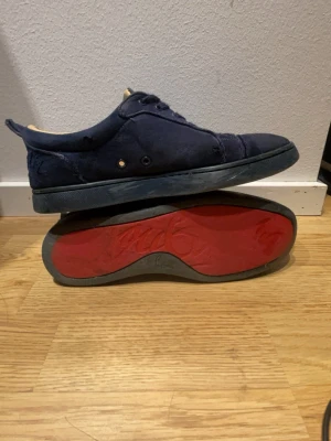 Mörkblå sneakers med röd sula - Snygga mörkblå sneakers med klassisk låg profil och röd sula. Skorna har ovandel i mocka, matchande mörkblå snören och metallöljetter. Insidan är beige och sulan har ett unikt mönster. Perfekta för dig som gillar stilrena detaljer.