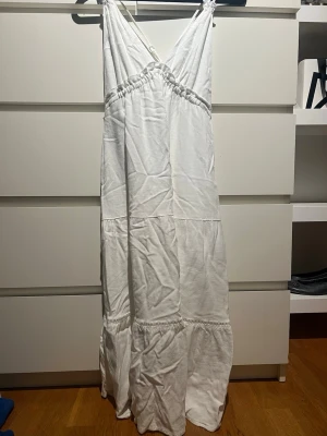 Vit långklänning med omlott och smala axelband - Vit långklänning med omlott framtill och smala korsade axelband. Klänningen har volangdetaljer och är tillverkad i ett lätt tyg, perfekt för varma dagar. Den har en luftig passform och en enkel, stilren look. Väldigt snygg passar perfekt till skolavslutning eller andra tillfällen.