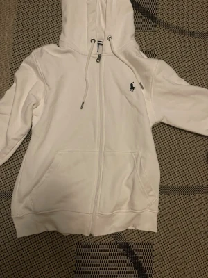 Vit hoodie från Ralph Lauren - Vit hoodie med dragkedja från Ralph Lauren. Klassisk design med huva och dragsko, två fickor framtill och den ikoniska broderade loggan på bröstet. Perfekt för en avslappnad och stilren look.
