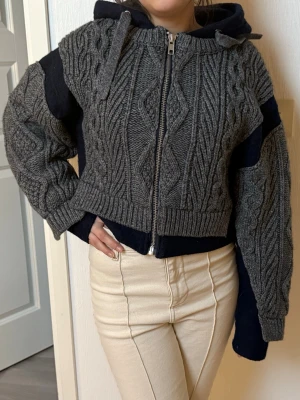 Grå stickad hoodie med dragkedja - Säljer en grå stickad hoodie med dragkedja framtill och huva med snören. Tröjan har ett grovt kabelstickat mönster och ribbade muddar. Ärmarna är vida och plagget har en croppad passform. Perfekt för dig som gillar en avslappnad men trendig Stockholm stil.