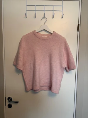 Ljusrosa stickad t-shirt - Söt ljusrosa stickad t-shirt med korta ärmar och rund hals. Tröjan har en avslappnad passform och ribbade kanter vid hals, ärmslut och nederkant. Perfekt för lager på lager och en mjuk känsla.