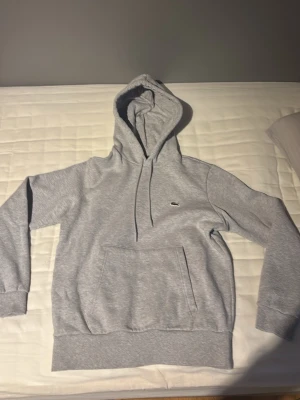 Grå hoodie från Lacoste med logga - Säljer en stilren grå hoodie från Lacoste med den klassiska krokodilloggan på bröstet. Hoodien har huva med dragsko, stor magficka och ribbade muddar vid ärmar och nederkant. Tillverkad i mjukt bomullsmaterial som är skönt att ha på sig.