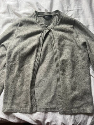 Grå kofta från Gina Tricot - Mysig grå kofta från Gina Tricot med lång ärm och ribbade kanter. Koftan har en enkel knapp framtill vid halsen och är öppen nedtill, vilket ger en avslappnad look. Perfekt att slänga över en topp för extra lager.