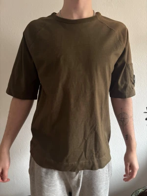 Olivgrön t-shirt från C.P. Company - Säljer en olivgrön t-shirt från C.P. Company med avslappnad passform och halvlånga ärmar. Tröjan har en ikonisk linsdetalj på ärmen och är gjord i mjuk bomull.
