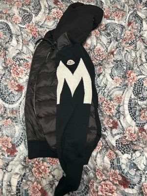  Moncler cardigan - Säljer en svart Cardigan  från Moncler med stor vit M-detalj på ärmen och klassisk Moncler-logga. Jackan har huva, dragkedja framtill och ribbade muddar. Perfekt för vår dagar och riktigt snygg streetstil.