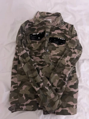 Camo overshirt med paljettdetaljer - Snygg overshirt i camouflage-mönster med svarta paljettdetaljer på ton anime-bröstfickor. Skjortkrage, knappstängning framtill och långa ärmar. Perfekt för dig som vill sticka ut lite extra.