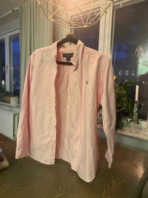 Ljusrosa skjorta från Ralph Lauren - Klassisk ljusrosa skjorta från Ralph Lauren med broderad logga på bröstet. Skjortan har knappar framtill, krage och långa ärmar. Tillverkad i mjuk bomull som känns skön mot huden. Perfekt för en stilren och avslappnad look.