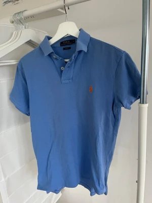 Blå pikéskjorta från Polo Ralph Lauren - Snygg ljusblå pikéskjorta från Polo Ralph Lauren i storlek S, custom fit. Klassisk krage, korta ärmar och två knappar framtill. Ikonisk orange logga broderad på bröstet. Skjortan är gjord i mjuk bomull som känns skön mot huden. Hör av dig om du har några frågor.