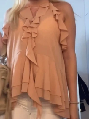 Beige volangblus med omlottdetalj - Säljer en beige blus med volanger och omlottdetalj framtill. Toppen är ärmlös och har en lös passform med snygga lager på framsidan. Perfekt för dig som gillar feminina detaljer och en luftig känsla.