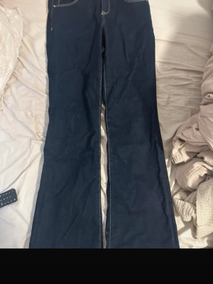 Mörkblå bootcut jeans med kontrastsömmar - Säljer ett par mörkblå bootcut jeans med vita kontrastsömmar och dubbla knappar i soppen. Klassisk femficksmodell i kraftigt denimtyg. Perfekta för dig som gillar en retroinspirerad look och vill ha ett par jeans med lite vidare ben.