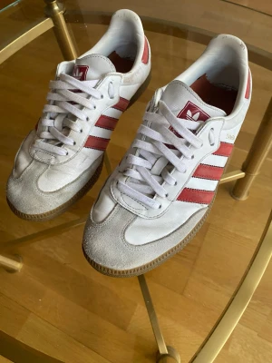 Adidas Samba strl 42 - Säljer ett par klassiska Adidas Samba sneakers i vitt skinn med röda detaljer och de ikoniska tre ränderna på sidan. De är i mycket bra skick bara att innersulans är utbytt.