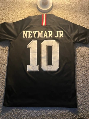 Neymar Jr 10 matchtröja svart PSG -  characteristisk svart PSG matchtröja med Neymar Jr och nummer 10 på inclusion. Framsidan har Fly Emirates-logga, PSG-emblem och Jordan-logga. Röd och vit rand på kragen och vertikal rand på bröstet. Perfekt för fotbollsfans.