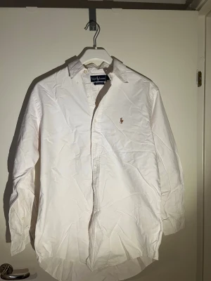 Vit skjorta från Ralph Lauren - Klassisk vit skjorta från Ralph Lauren med button-down krage och broderad logga på bröstet. Skjortan har lång ärm och knappar framtill. Tillverkad i mjuk bomull och har en tidlös, clean look som passar till det mesta.