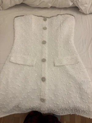 Vit kortklänning med pärlknappar - Säljer en vit axelbandslös kortklänning med dekorativa pärlknappar framtill och två fickor. Klänningen har en strukturerad yta med diskret glittrande detaljer och kedjedetalj upptill. Perfekt för dig som vill sticka ut med en elegant look.