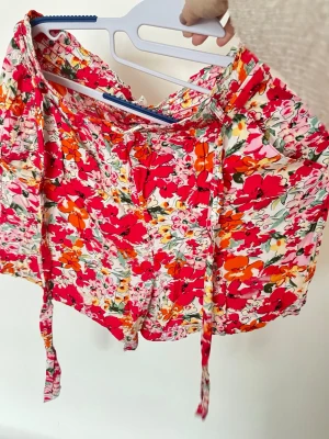 Blommiga shorts med knytband - Färgglada shorts med blommönster i rött, orange, rosa och grönt på vit botten. Modellen har bred resår i midjan och långa knytband framtill. Luftigt material, perfekt för sommaren. Passar dig som gillar en lekfull och trendig stil.