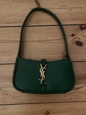 Ysl väska  - Snygg mörkgrön handväska från Saint Laurent i slät läder med guldfärgad YSL-logga framtill. Väskan har en klassisk axelrem och dragkedja upptill. Insidan är fodrad i vinrött tyg och har märkning med Saint Laurent Paris. Perfekt accessoar för en trendig look.