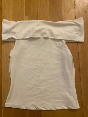 Vit offshoulder topp från H&M - Säljer en stilren vit offshoulder topp från H&M. Toppen är axelbandslös och har en tight passform som framhäver figuren. Perfekt att matcha med både jeans och kjol för en trendig look. Materialet IMITerar bomull och känns mjukt mot huden.