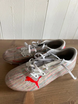 s/Gråa Puma fotboll stained skor - Säljer ett par gr sopade fotbollsskor från Puma med vita snören och röda Puma-loggor på ovansidan. Skorna har ett randigt mönster i grått, vitt och rött och är tillverkade i syntetmaterial för bra grepp och lätt känsla på planen.