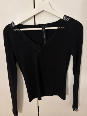 Svart långärmad topp med spets - Svart långärmad topp från The Black Label Collection med v-ringning och dekorativ spetskant runt halsen. Tunn och stretchig modell som sitter snyggt på kroppen. Perfekt att styla med jeans eller kjol.