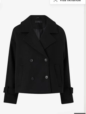 Svart kort dubbelknäppt rock från Ellos - Svart kort rock från Ellos med dubbelknäppning och breda slag. Rocken har två framfickor och justerbara ärmslut med knappar. Materialet ser ut att vara ullblandning och den har en klassisk, boxig passform som är perfekt för kalla dagar.