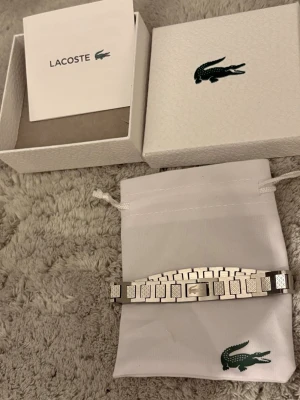Silverfärgat armband från Lacoste - Stilrent armband i silverfärgad metall från Lacoste med rektangulära länkar och den klassiska krokodil-loggan ingraverad på mitten. Kommer med original ask och tygpåse med Lacoste-logga. Perfekt accessoar för en modern look.